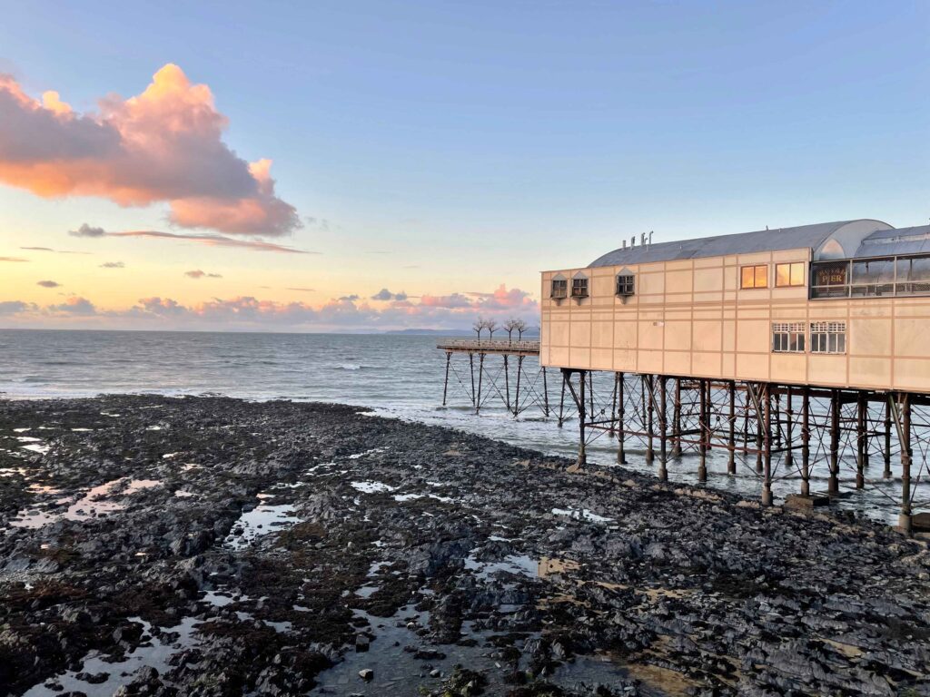 The rocky shoreline in Aberystwyth at low tide. The city's pier is an enclosed, two-story building. 