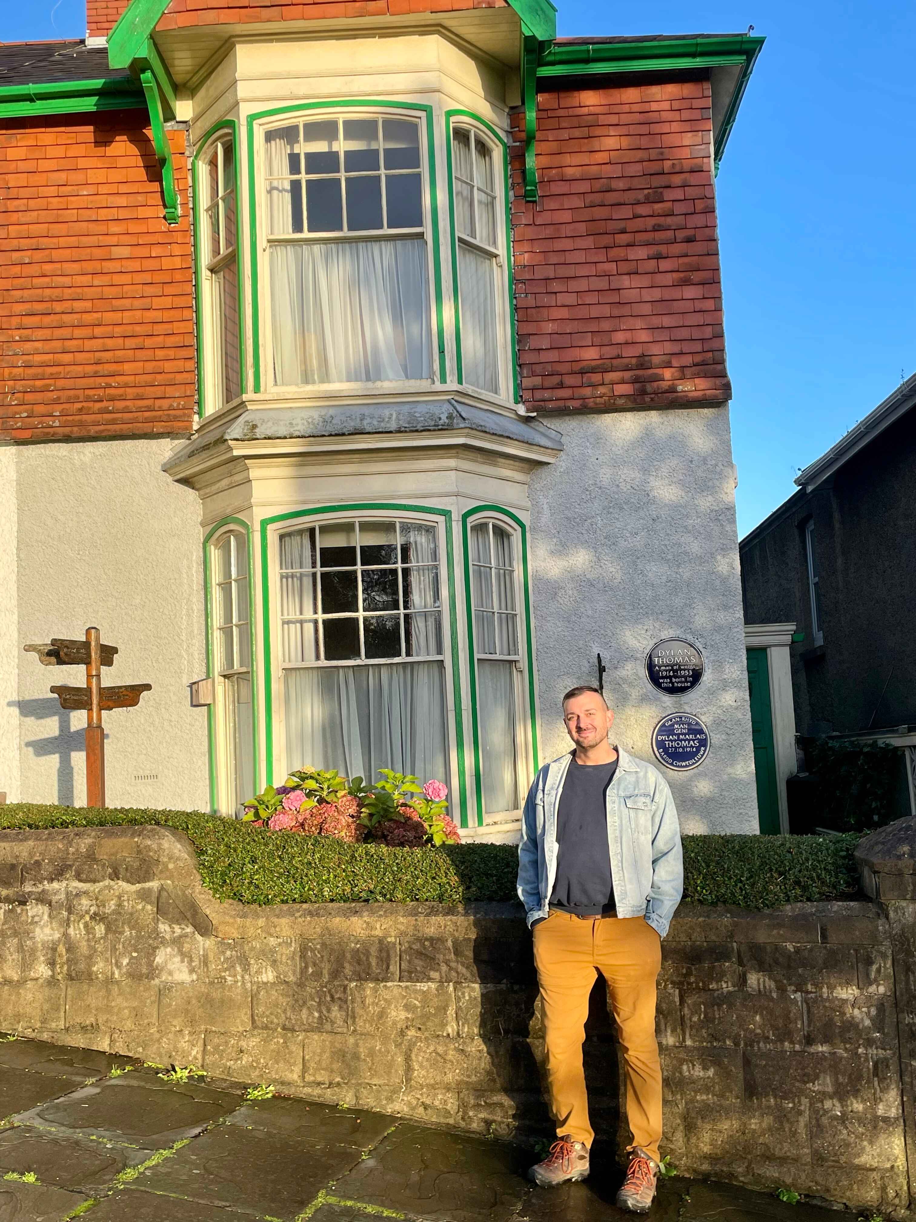 The house of poet and writer Dylan Thomas. A low brick wall, a neatly trimmed hedge. There is a bay window, and some placards on the wall of the house. 