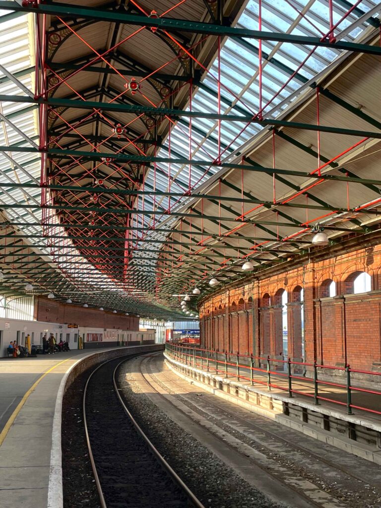 The interior of the Victorian era train station in Holyhead, Wales. Brick walls, and a single rail track with a few passengers waiting in the background. 