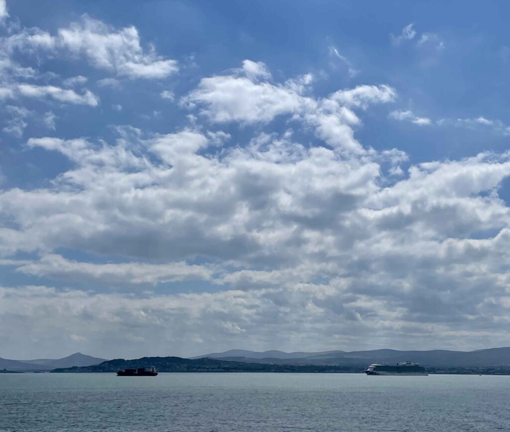 The coast of Ireland, with fluffy white clouds in the sky, low-lying mountains in the distance, and two boats in the ocean. 