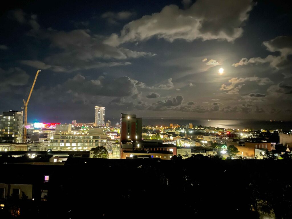 The city of Swansea seen from a hilltop at night. There is a full moon, a few clouds in the sky. There are a few tall apartment buildings, and a construction crane. 